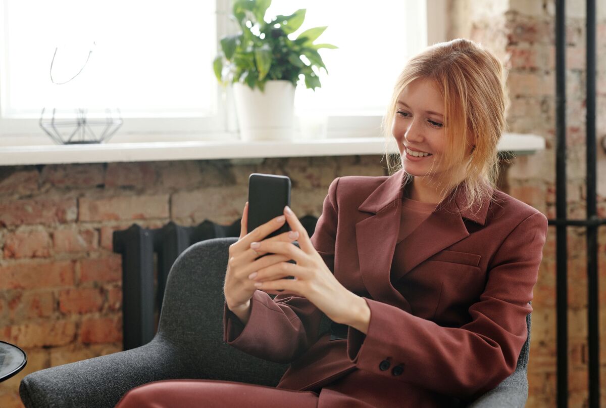 Eine Frau sitzt in einem Sessel und lächelt auf ihr Smartphone.